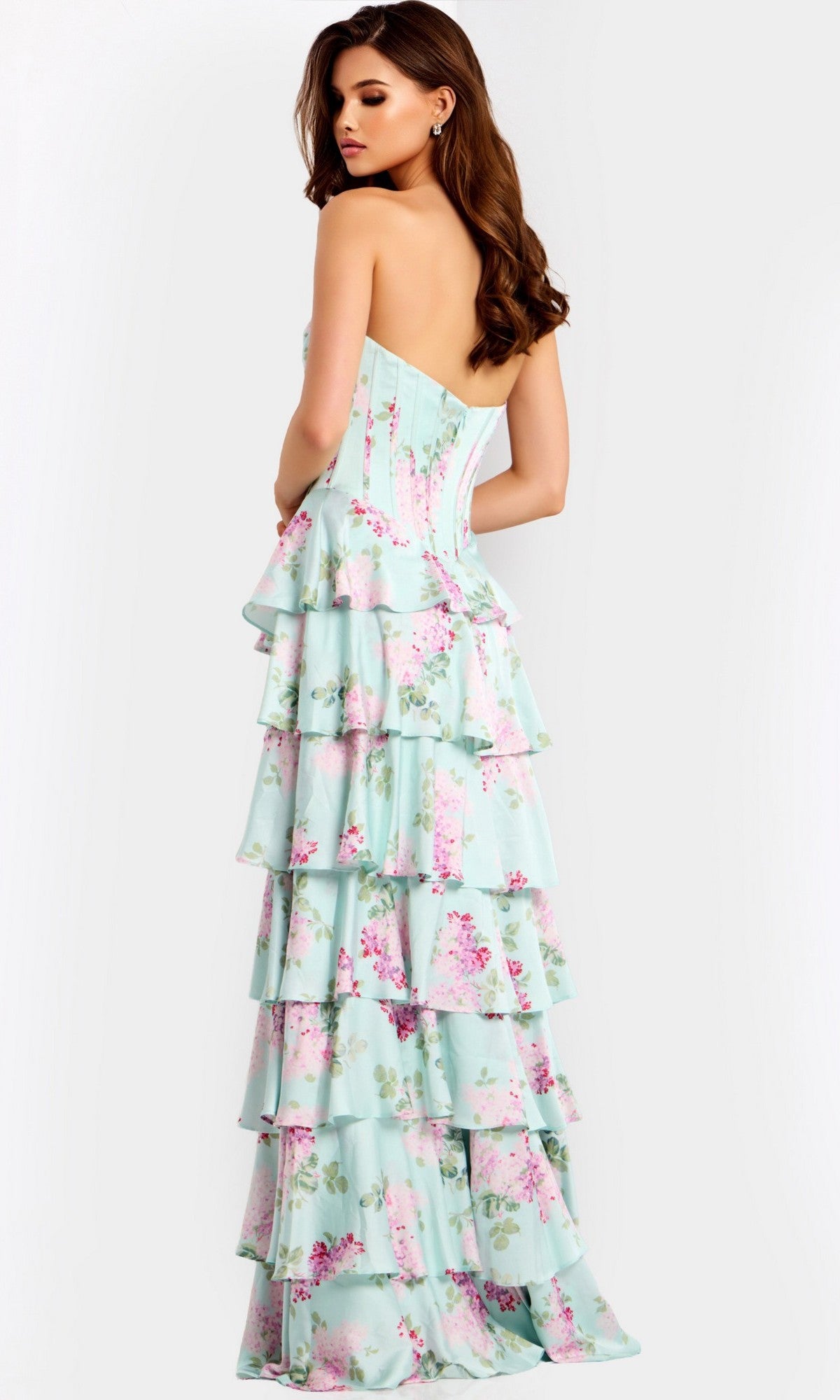 A woman stands with her back turned on a light gray background, wearing the Jovani 47579 long prom dress—a strapless gown in pastel blue with pink floral print, corset bodice detail, and tiered ruffles.